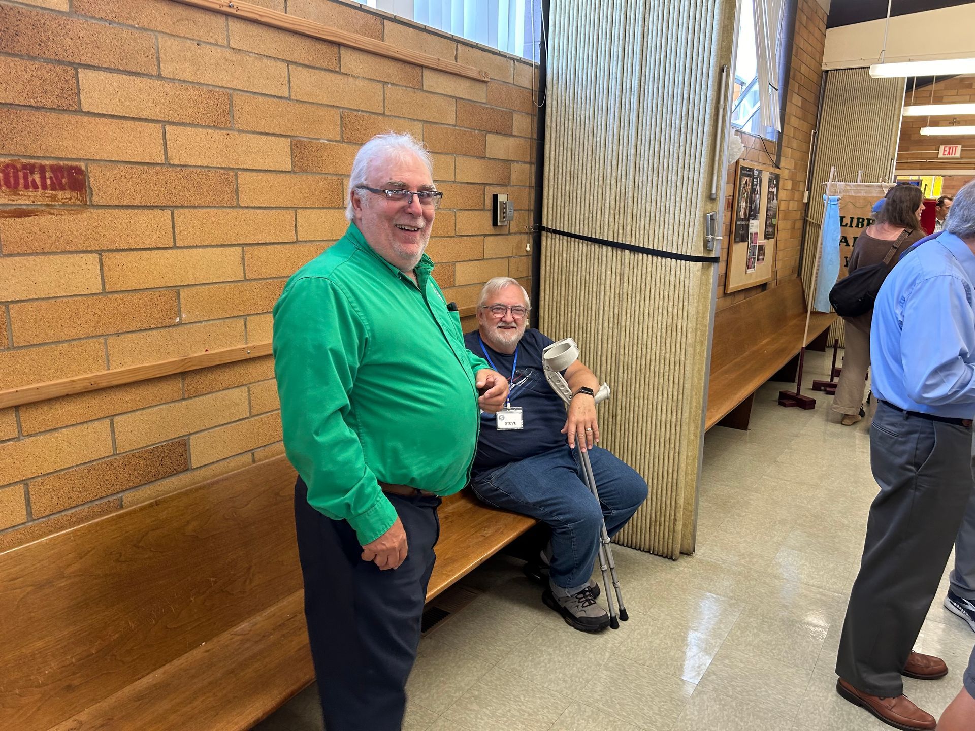 Two men in casual clothes sit and stand in a hallway by a wooden wall and brick wall.