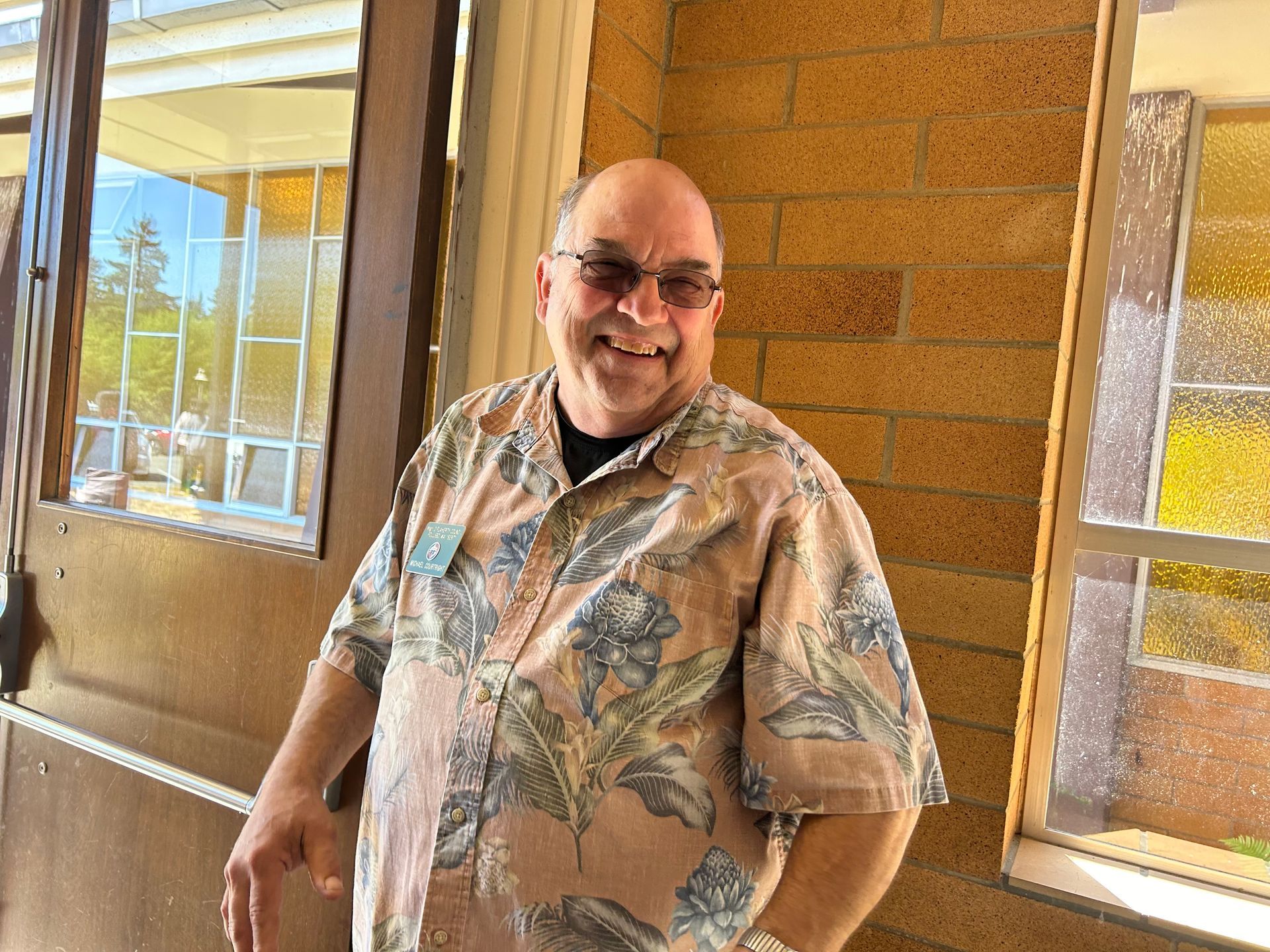 Mike smiling in a floral shirt standing by a windowed brick wall in a sunlit hallway