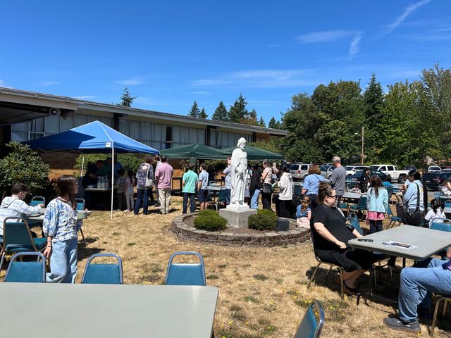 Outdoor gathering with people seated and standing around a white statue in front of a building, under blue skies