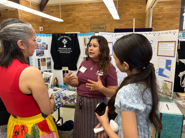 Three people talk at a poster display in a social hall.