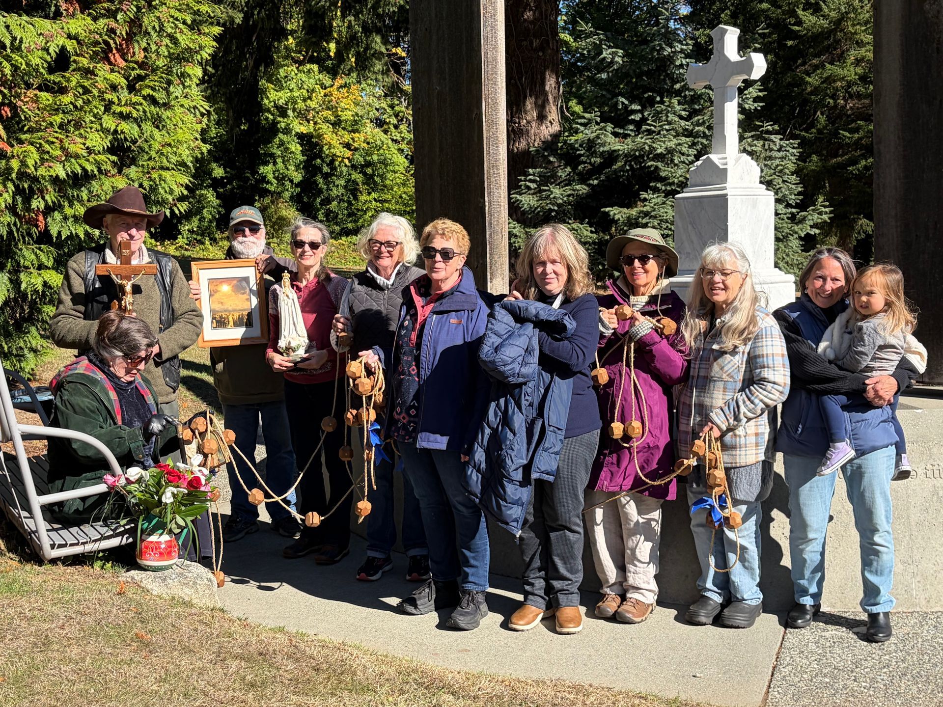 Group of people standing outdoors beside a white cross-shaped monument in a garden setting