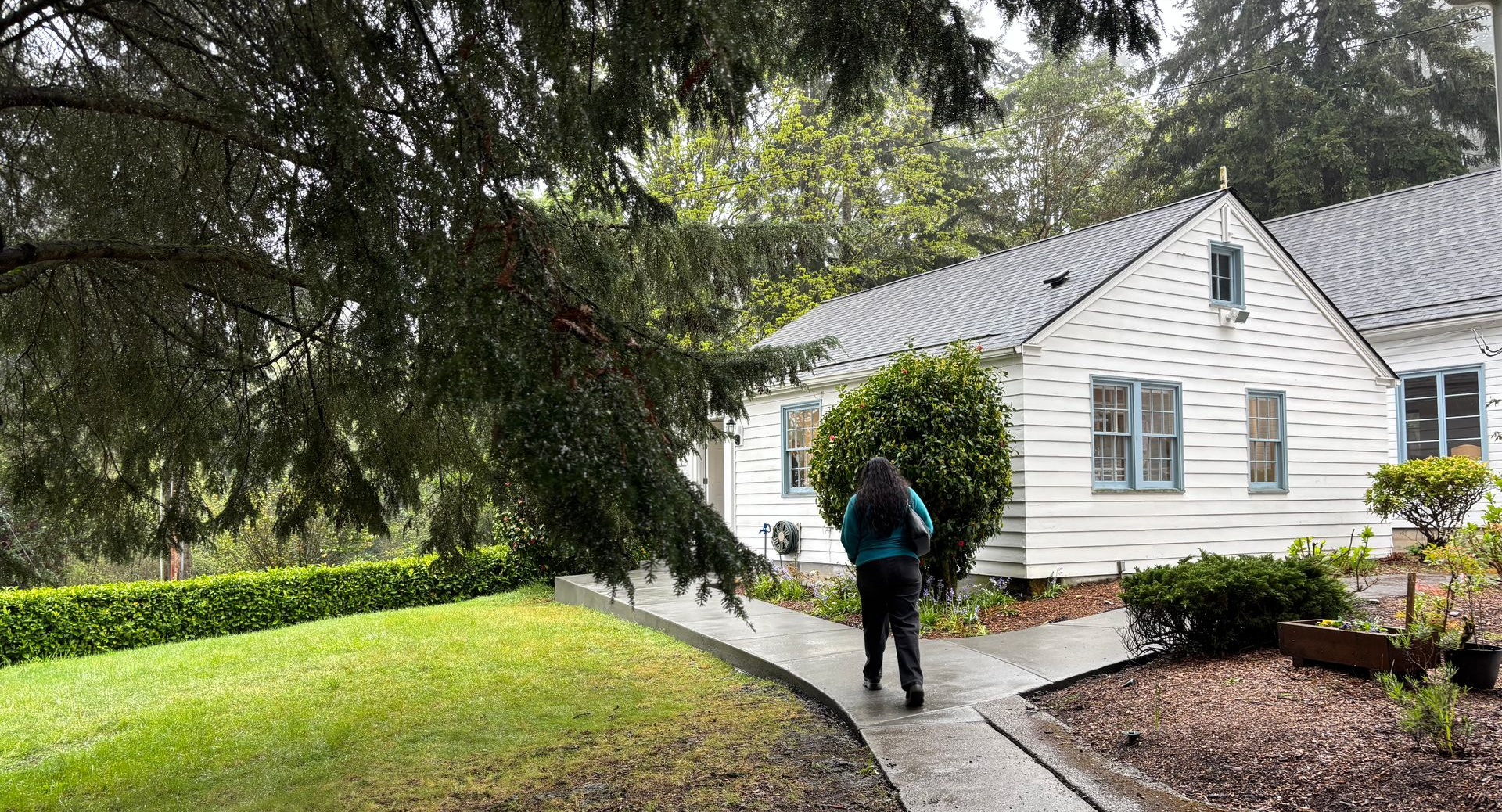 Person walking on a path toward a white house with a gray roof, surrounded by green landscaping