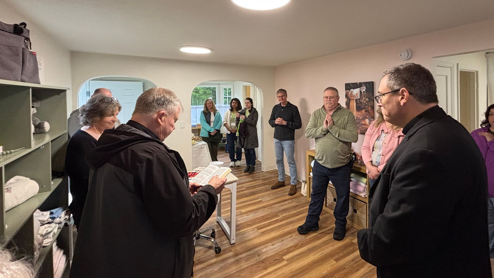 Group of people standing in a room during a meeting, listening to a speaker near shelves and wooden floors