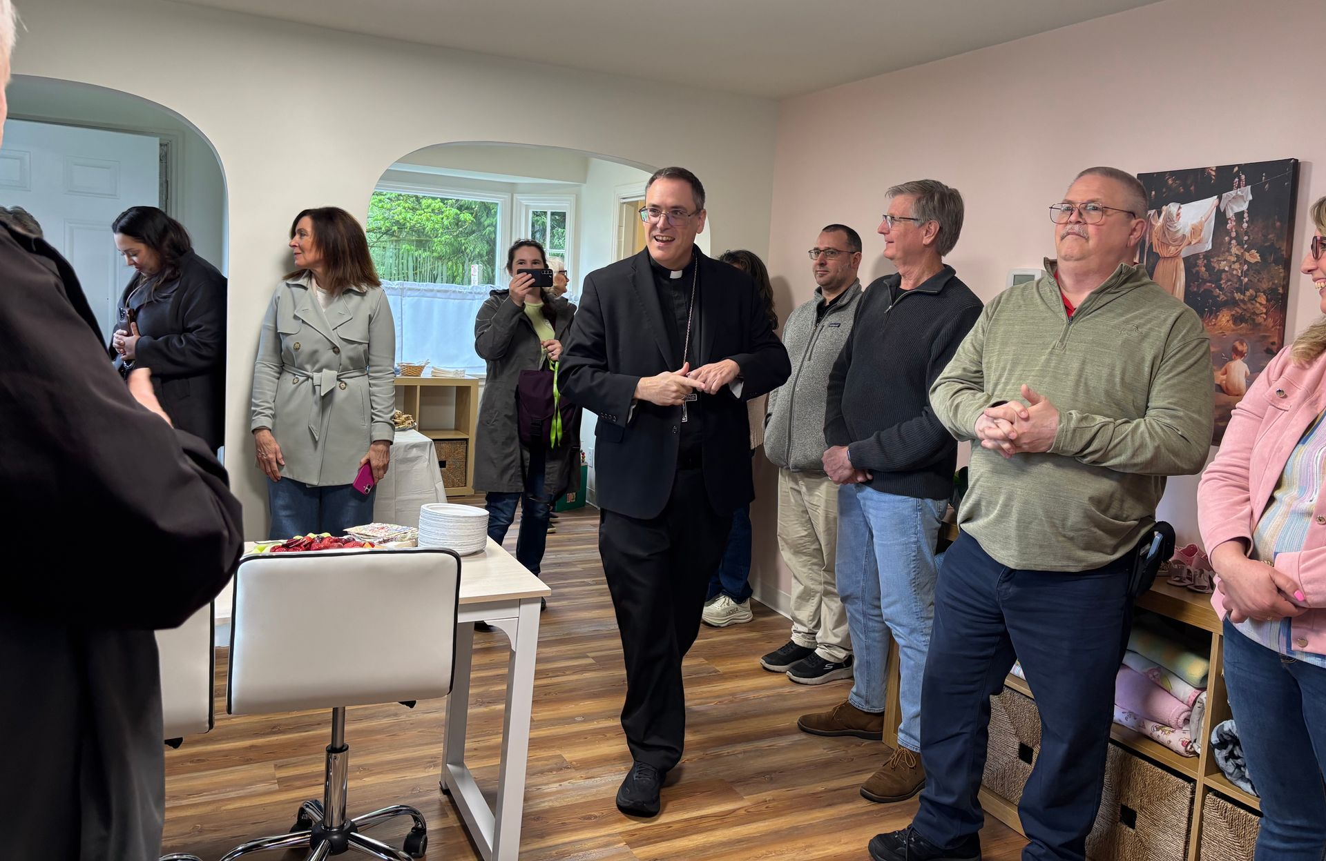 People standing and listening in a bright room during a small indoor gathering.