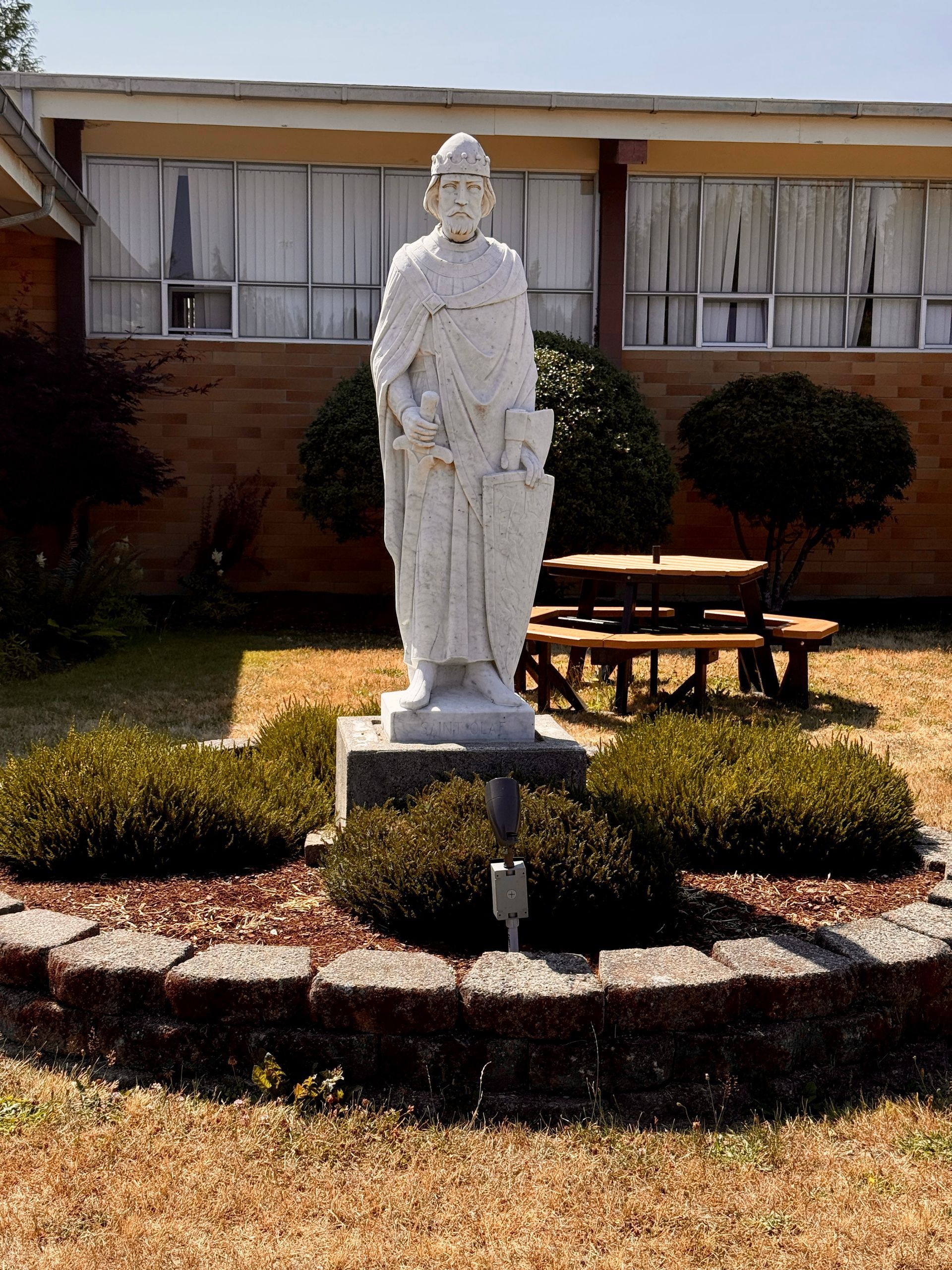 White statue of St. Olaf on a pedestal in a landscaped courtyard outside a building with windows and picnic tables.