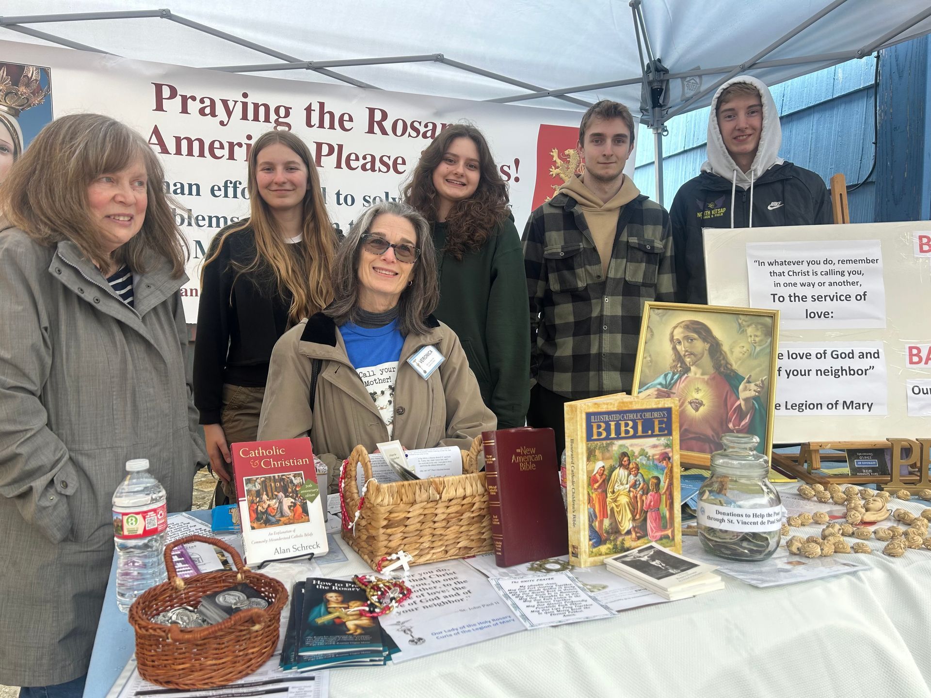 Booth with books and rosaries; five people stand behind a table under a “Praying the Rosary” banner.