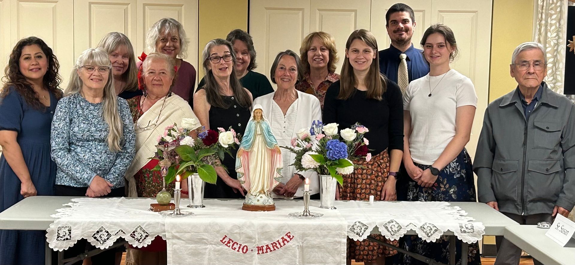 Group of people posing behind a decorated table with flowers for Mary in a bright indoor room.