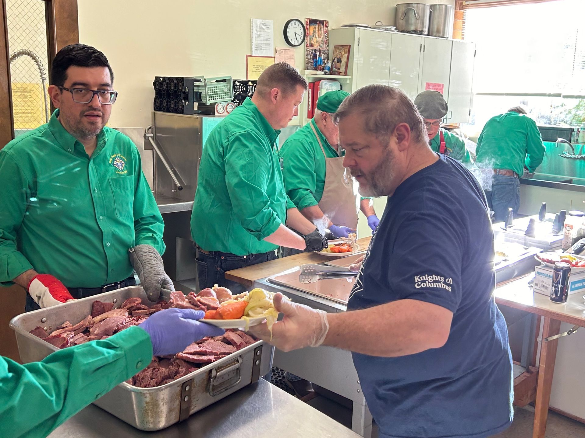 Workers in green uniforms prepare food in a kitchen, sorting trays of meat and vegetables.