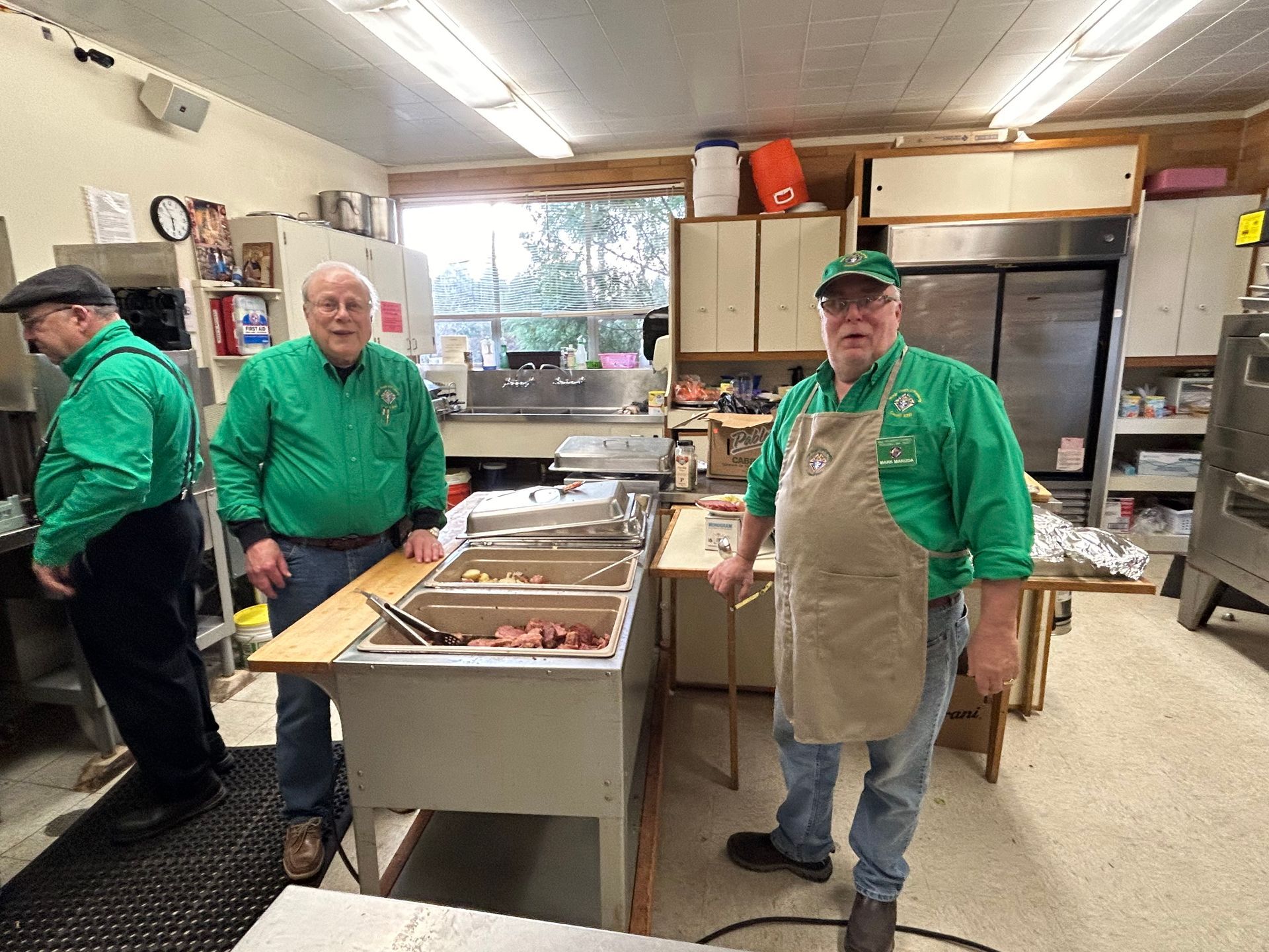 Men in green shirts and aprons stand in a kitchen with stainless steel prep tables.