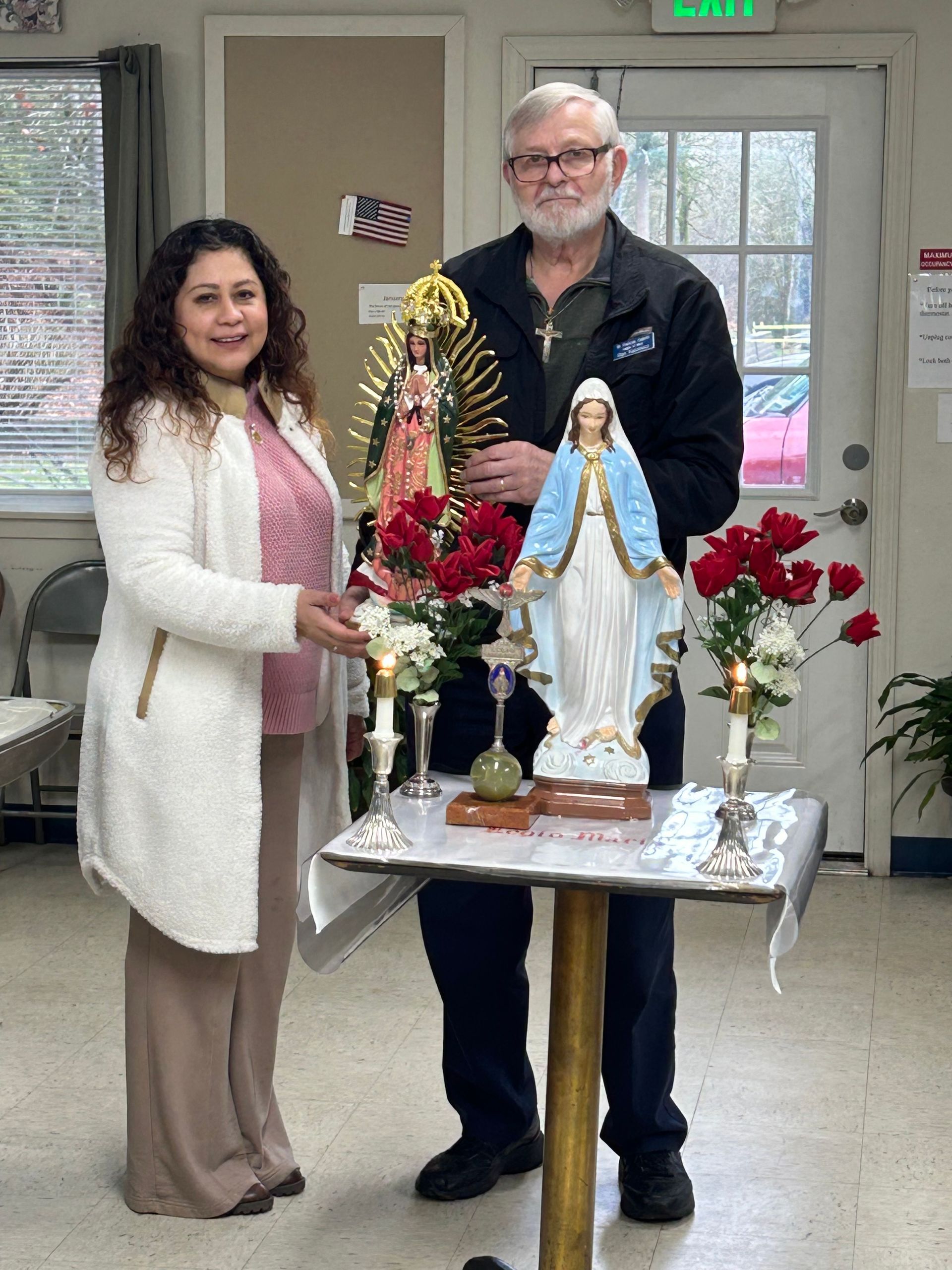 Two people standing behind a decorated table with religious statues and red flowers indoors.