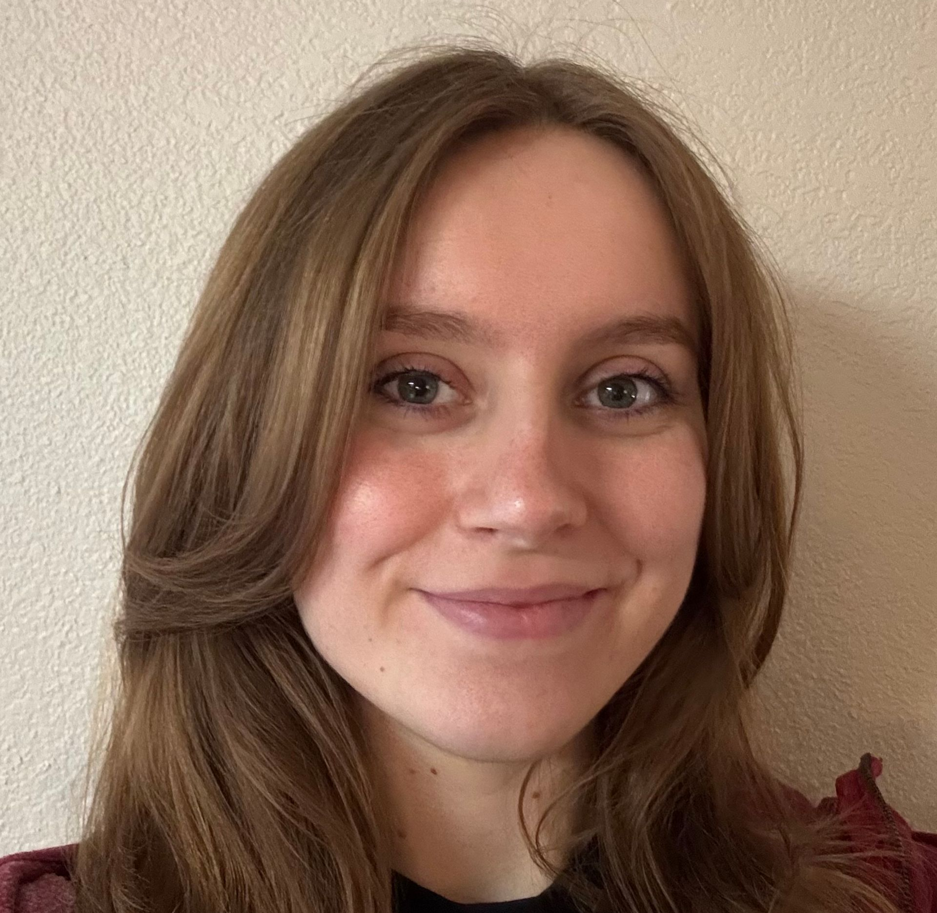 Maggie smiling with long brown hair against a beige wall