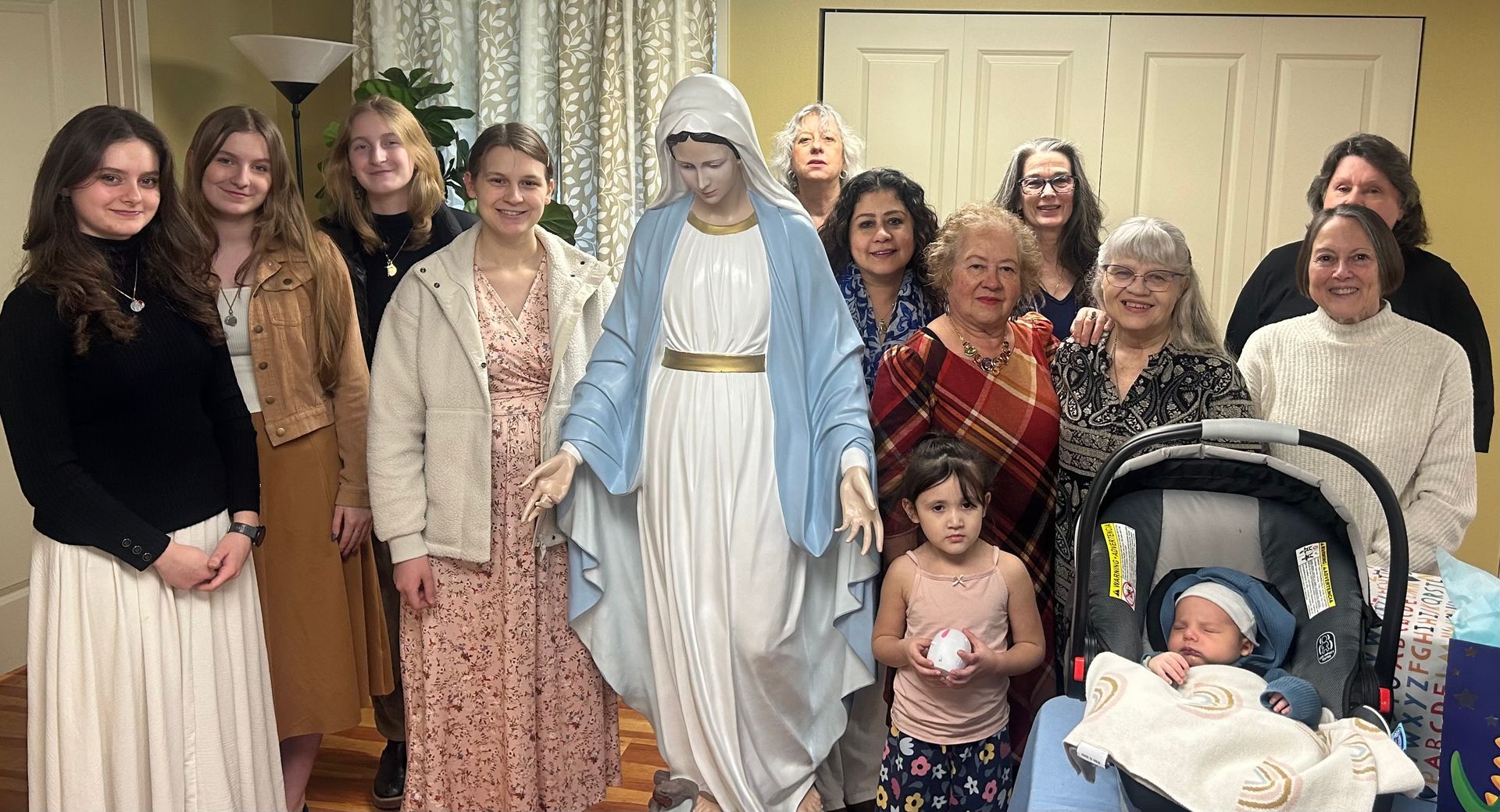 Group of women poses indoors beside a statue of Mary with a baby in a stroller.