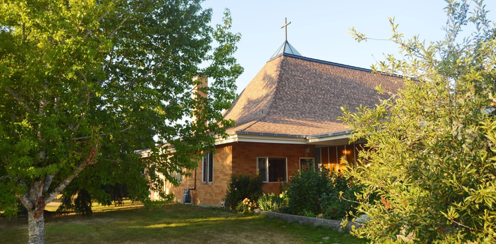 Back of St. Olaf Church with a cross on the roof, surrounded by green trees in warm sunlight.