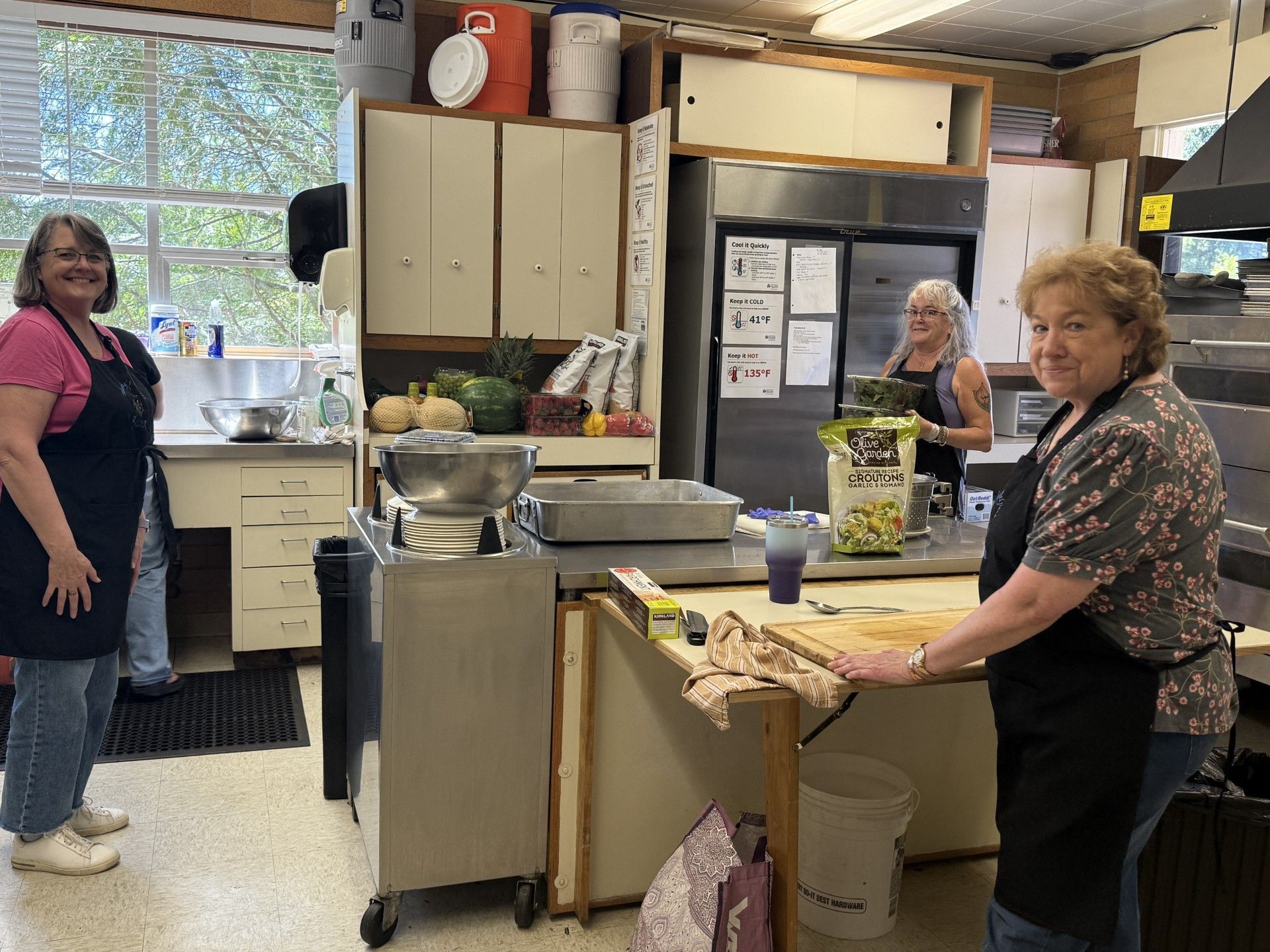 People in a kitchen with counters, cabinets, and pots; one woman stands by the sink, another leans on a counter.