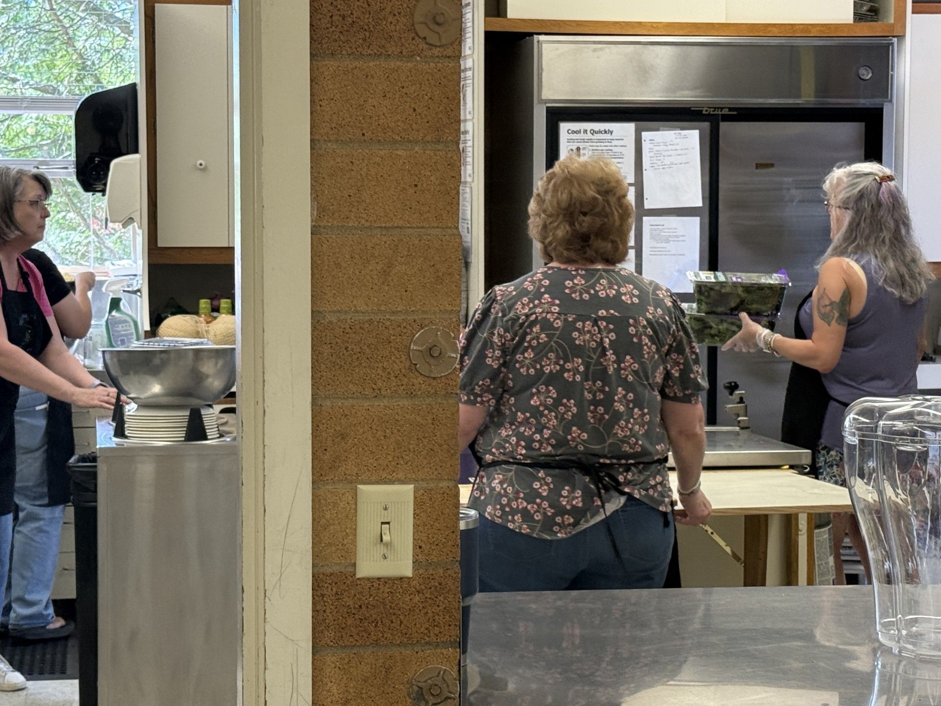 People standing at a serving counter in a cafeteria, with trays and a large bowl on the counter.