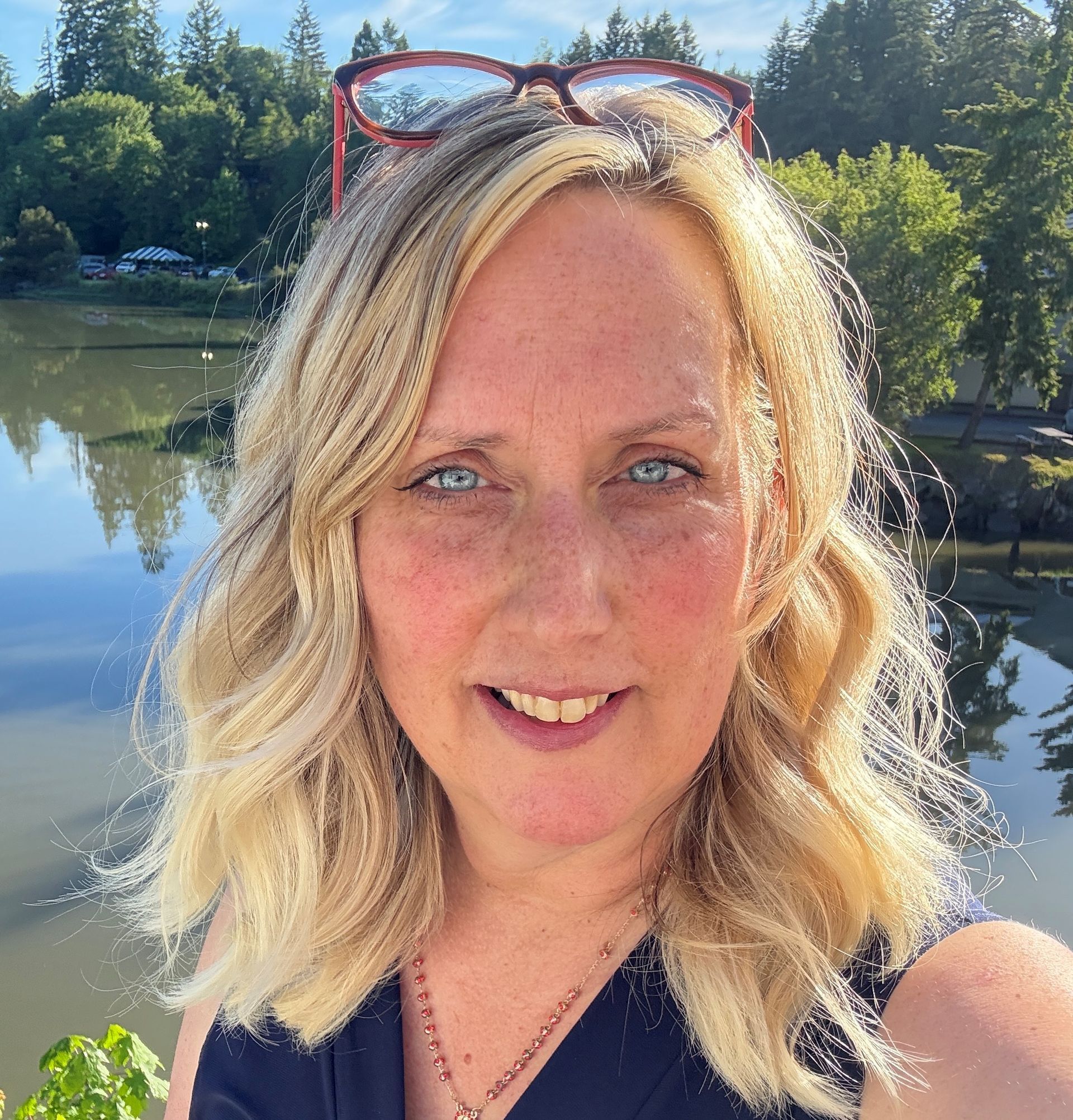 Valerie smiling outdoors by a calm lake, with glasses on her head and trees reflected in the water