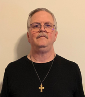 Kurt wearing glasses and a gold cross necklace, standing against a plain beige wall.