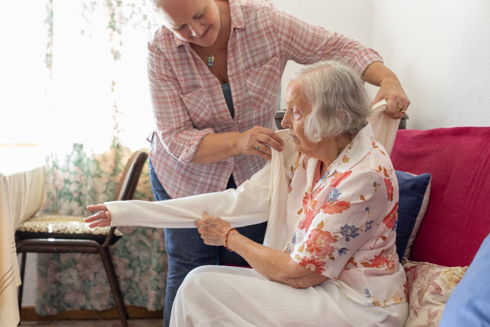 Woman helping elderly woman put on a cardigan indoors; both smiling. Woman helping elderly woman put on a cardigan indoors; both smiling.
