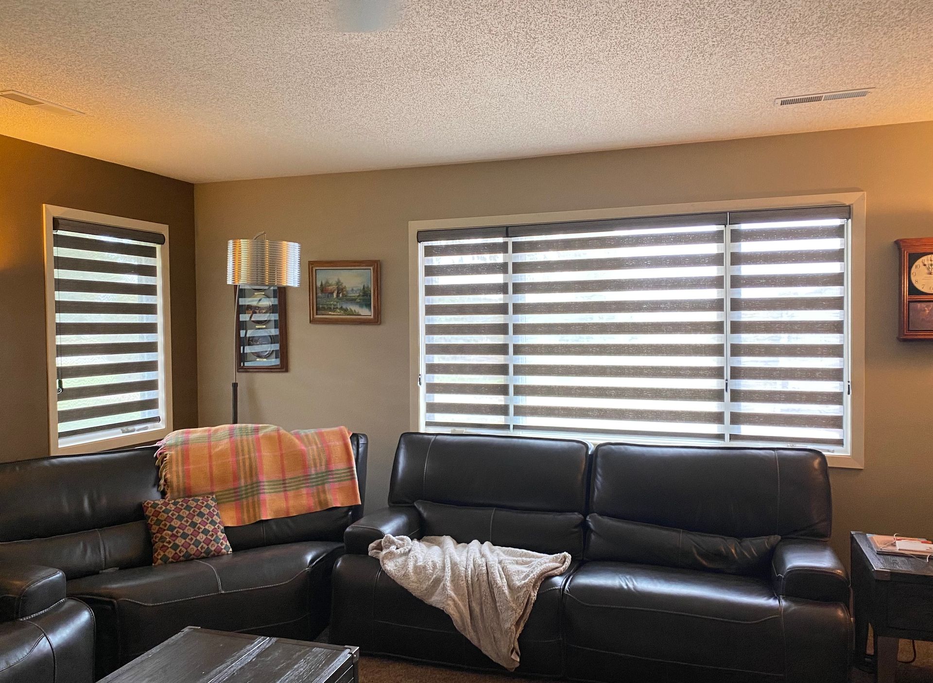 A living room with a black leather couch, dual-shade blinds, and a patterned blanket.