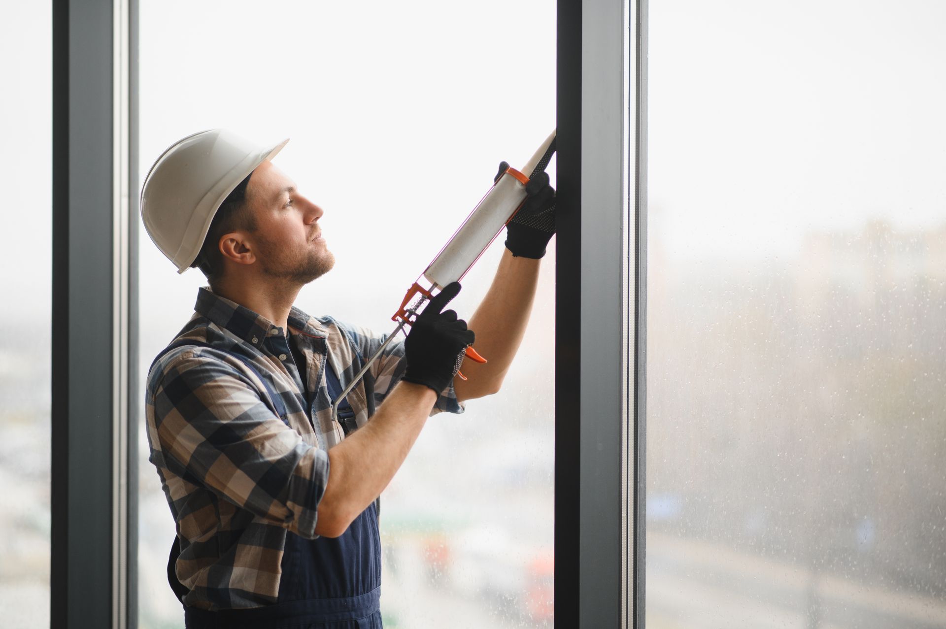 Worker applying sealant with caulking gun along window frame indoors.