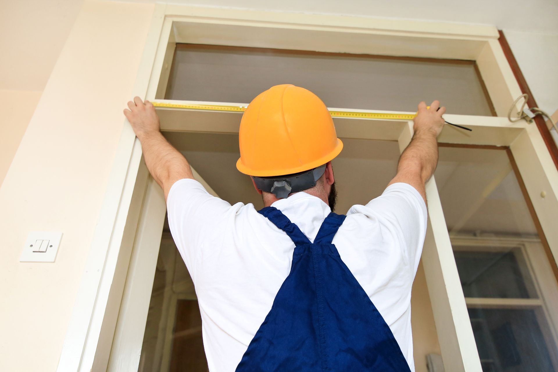 A man in a hard hat and overalls measures a door frame with a tape measure. A man in a hard hat and overalls measures a door frame with a tape measure.