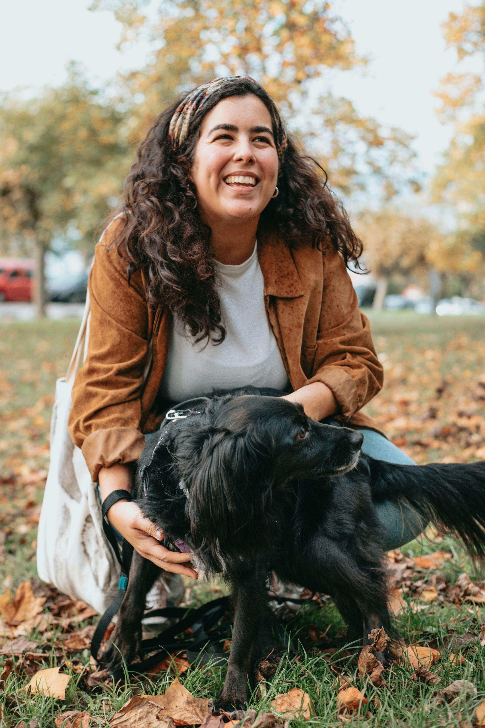 Woman with curly brown hair smiles while petting a black dog in a park with autumn leaves.