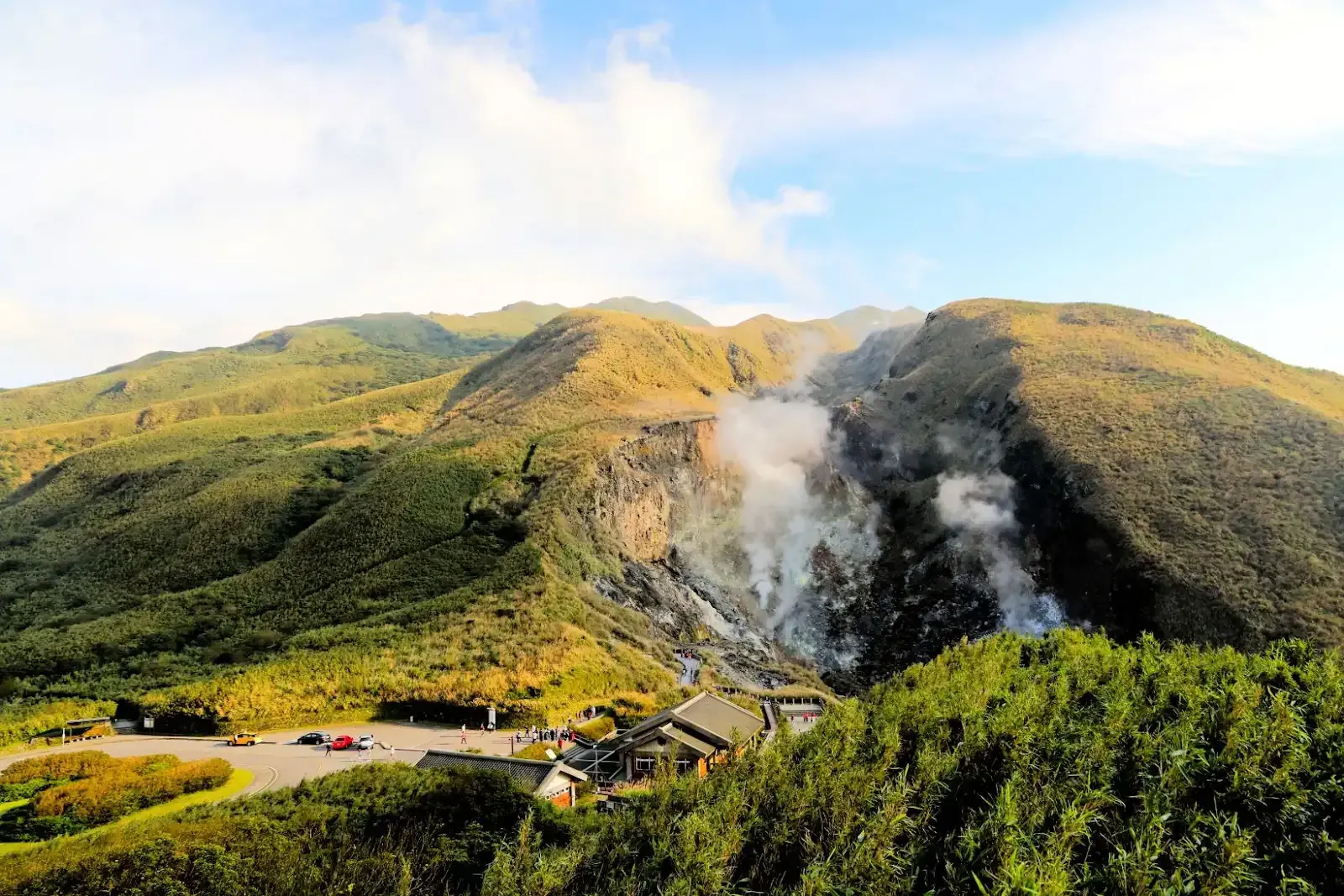 山巒蒸騰，蒸氣升騰，山腳下有建築物及車輛。山坡上綠樹成蔭，天空湛藍。