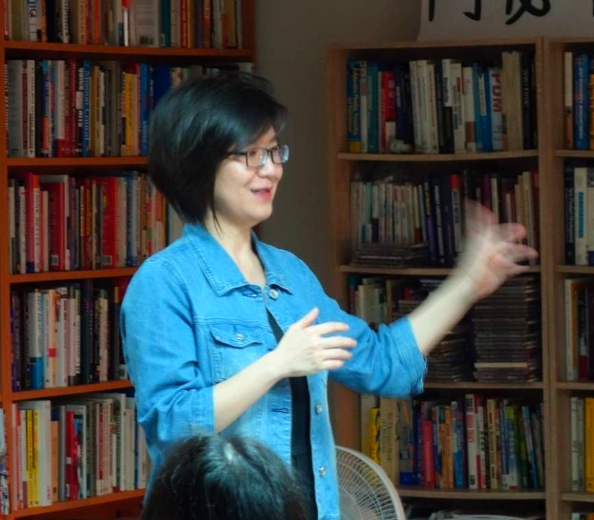 一位身穿藍色牛仔外套的女子在書架前熱情地做著手勢。