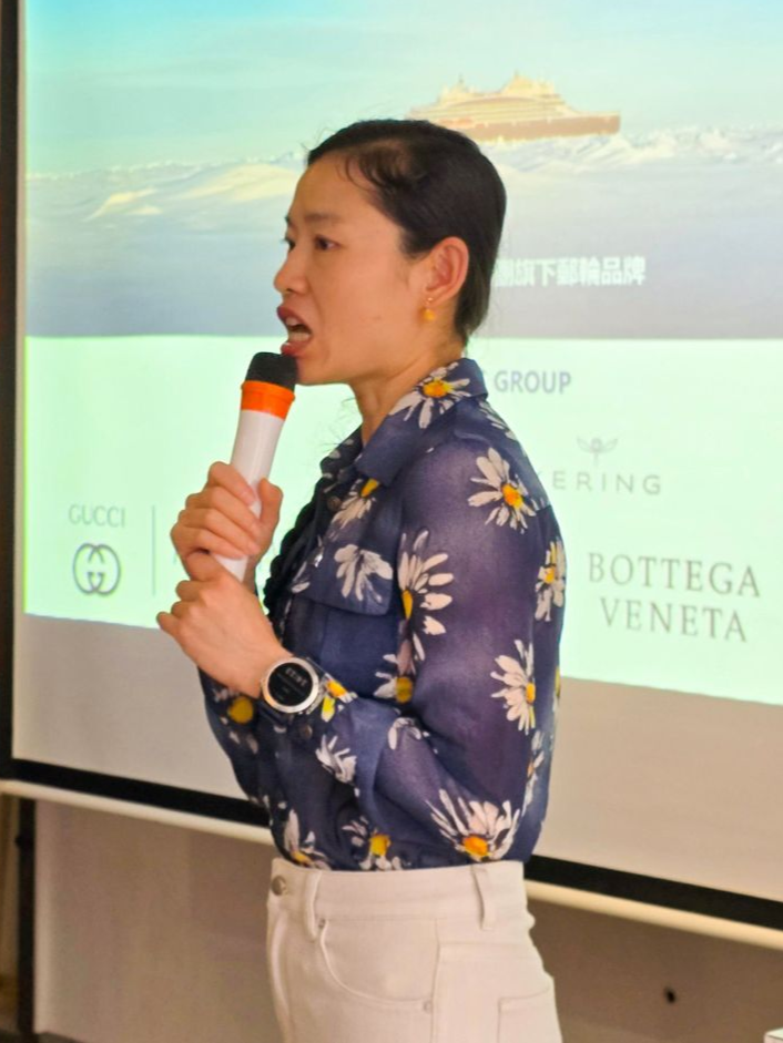 一名女子對著麥克風講話,她站在一個印有奢侈品牌標誌的螢幕前。