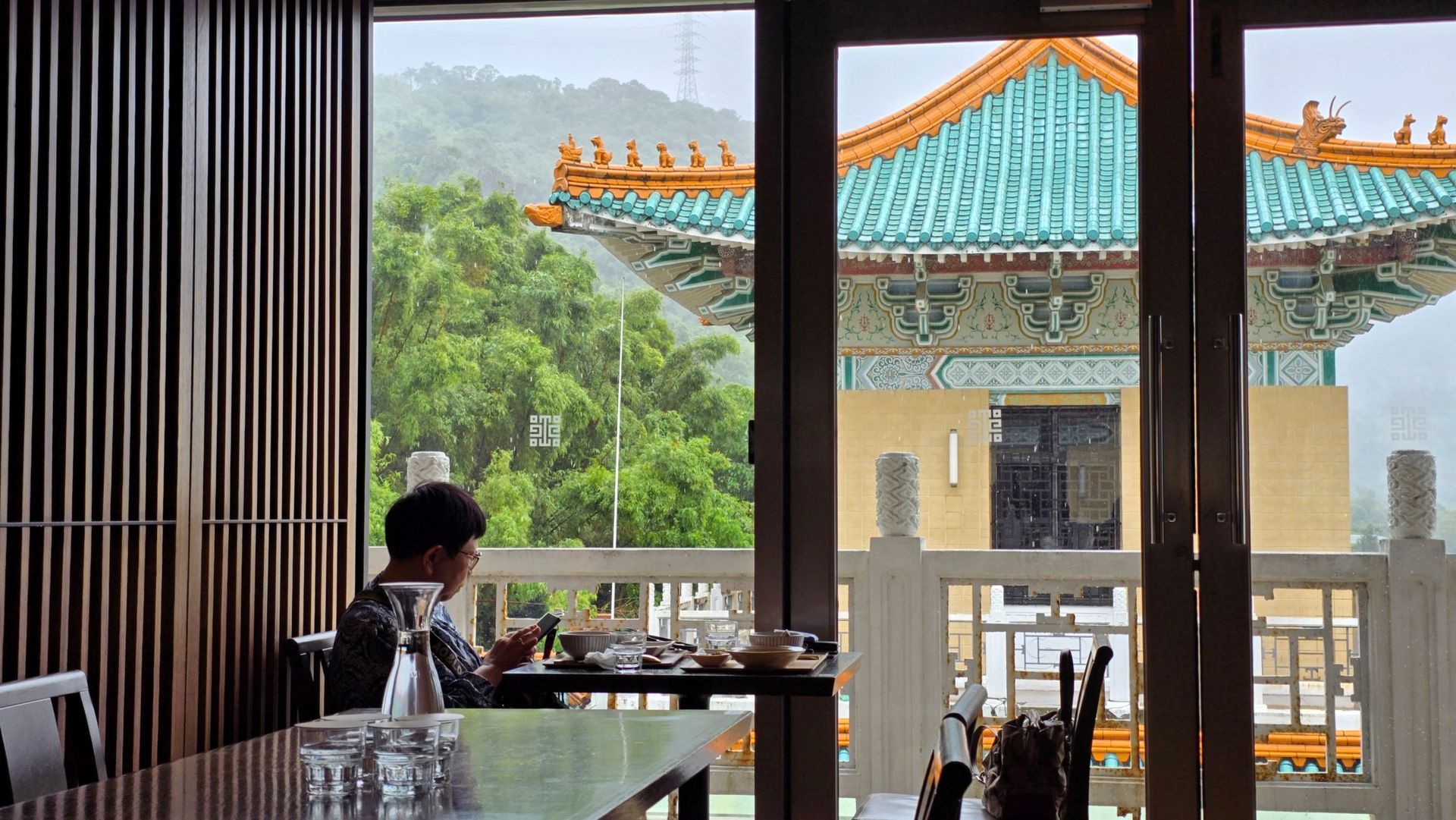 餐廳內，一位顧客坐在桌旁，望著一棟屋頂泛綠的建築，遠方是山巒。
