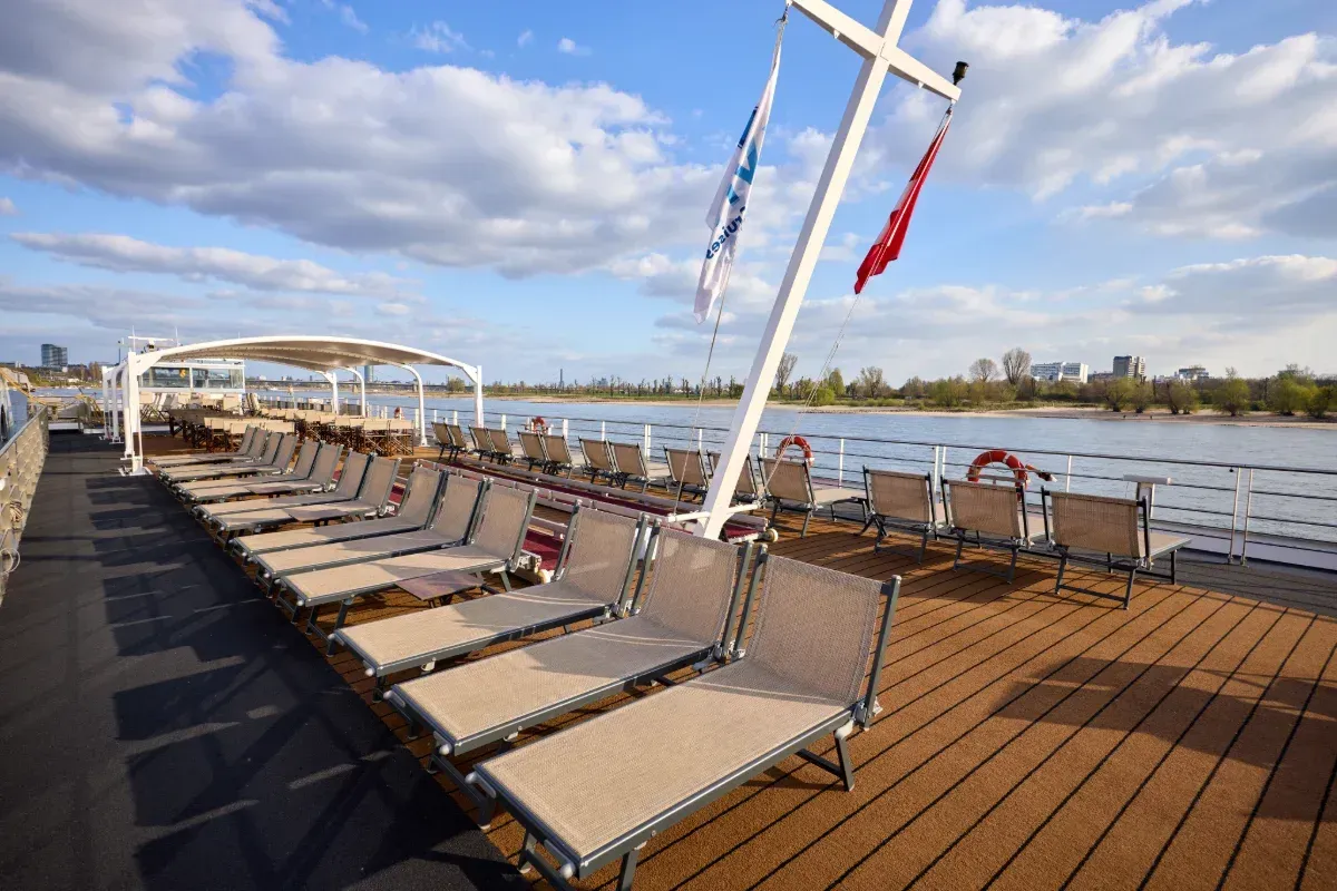 Lounge chairs on a wooden deck by a river, with cloudy sky and flags on poles.