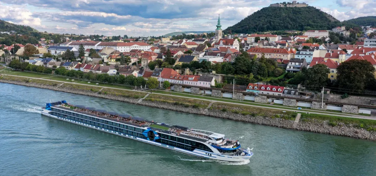 River cruise boat passes a riverside town beneath a green hill and cloudy sky