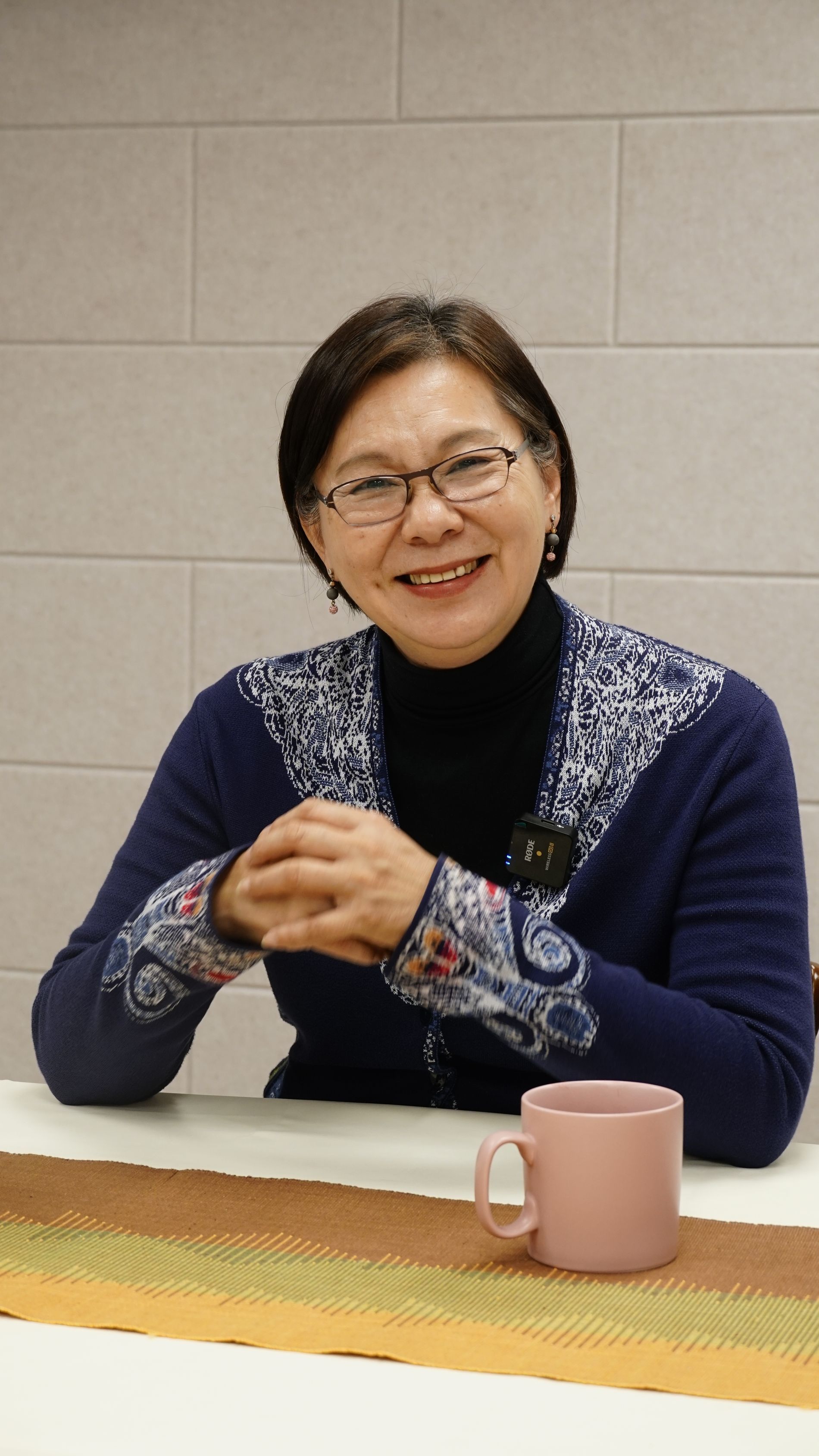 一位面帶微笑、戴著眼鏡、身穿藍色花紋開襟衫的女士坐在桌旁，桌上放著一個馬克杯。