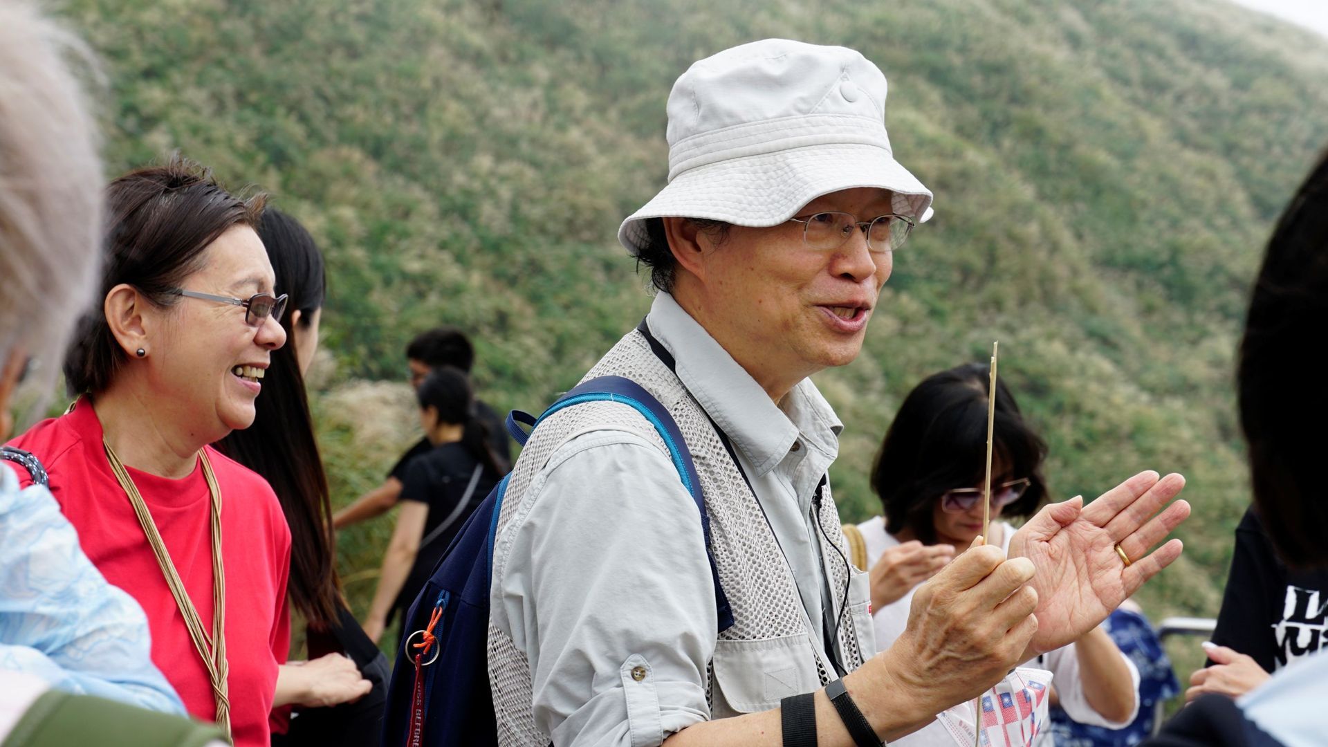 一名戴帽子的男子一邊做手勢一邊與一群人交談；他身旁的一名女子麵帶微笑。背景是連綿的山脈。