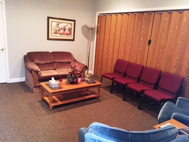 Small waiting room with brown couch, red chairs, coffee table, floor lamp, and wood-paneled wall.