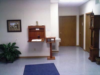 Quiet hallway with wall-mounted wooden shelving, potted plant, blue mat, and a grandfather clock.