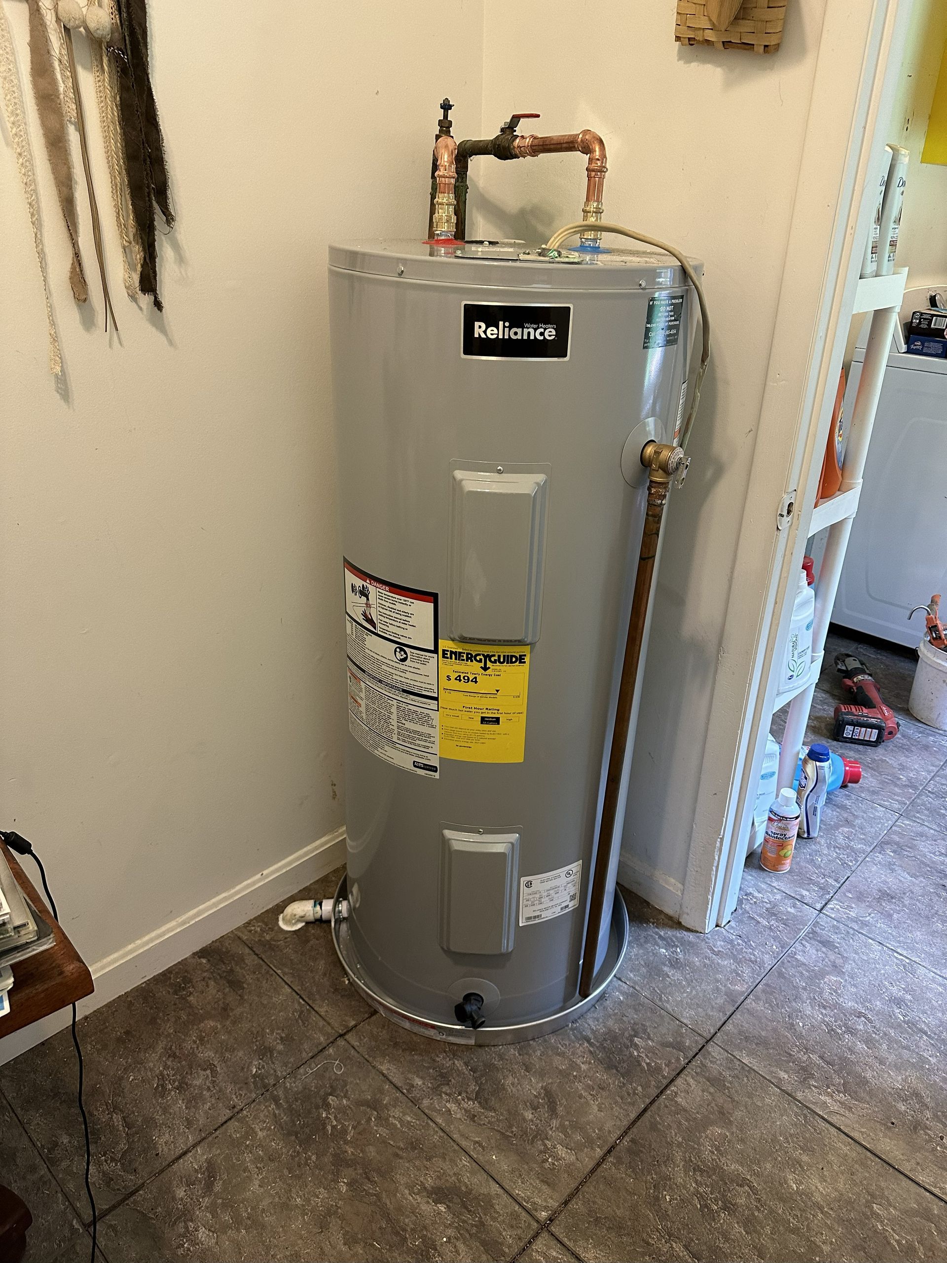 A tall, gray water heater in a corner, with copper pipes and yellow energy label.