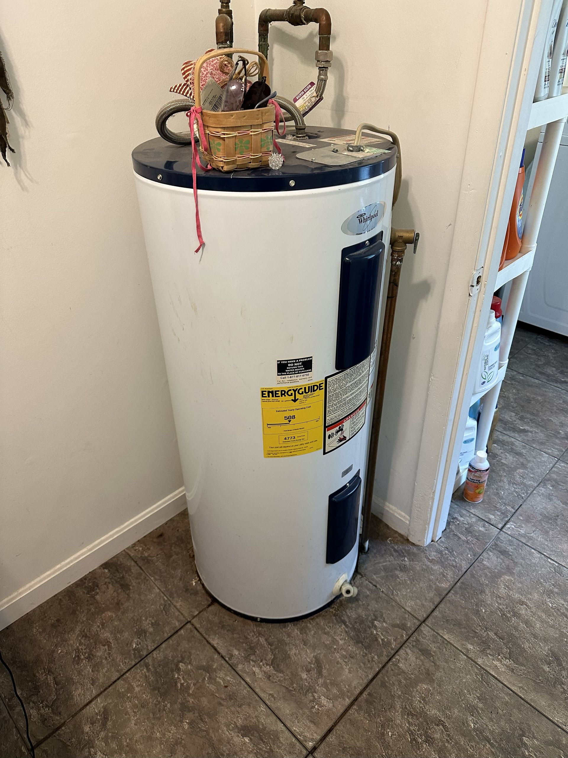 White water heater in a corner, with pipes and small items on top, set on tiled floor.