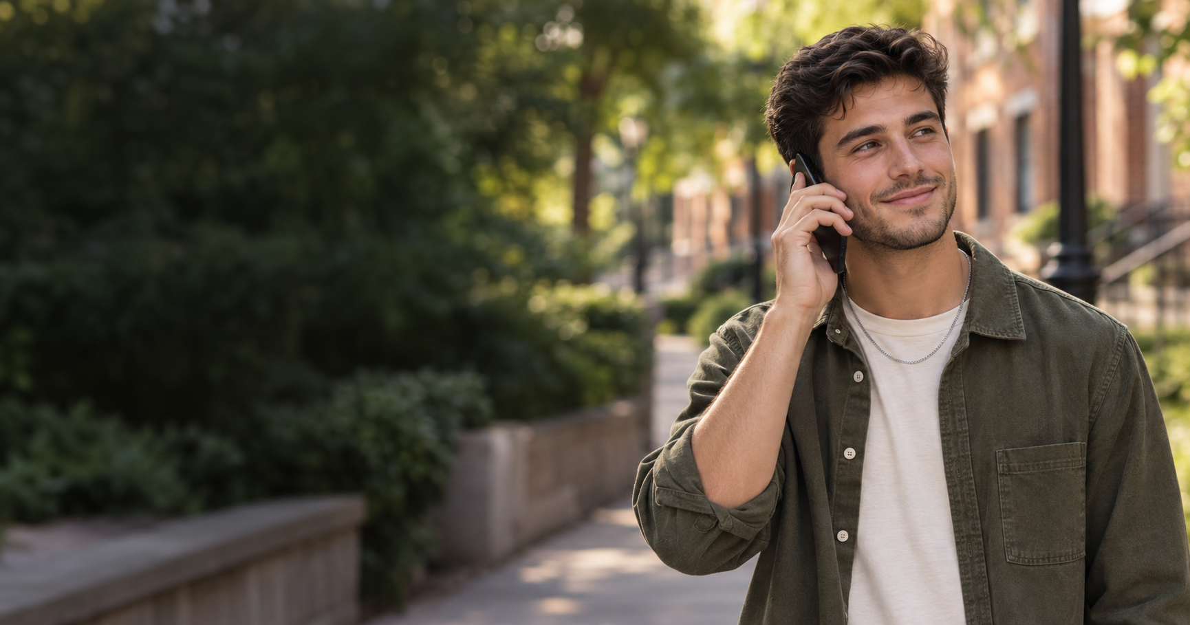 Smiling man in an olive shirt talking on a phone on a sunny city sidewalk