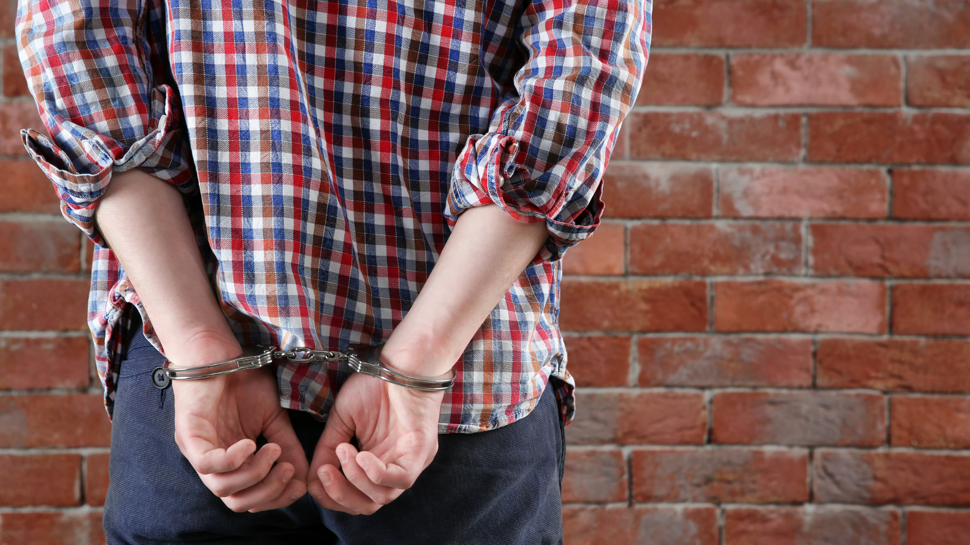 Handcuffed hands behind back against a brick wall, wearing a red and blue plaid shirt