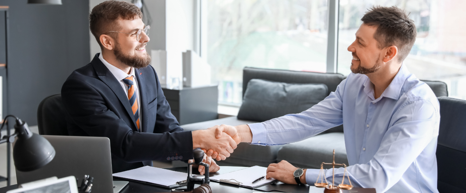 Two businessmen in suits shake hands across a desk in a modern office.