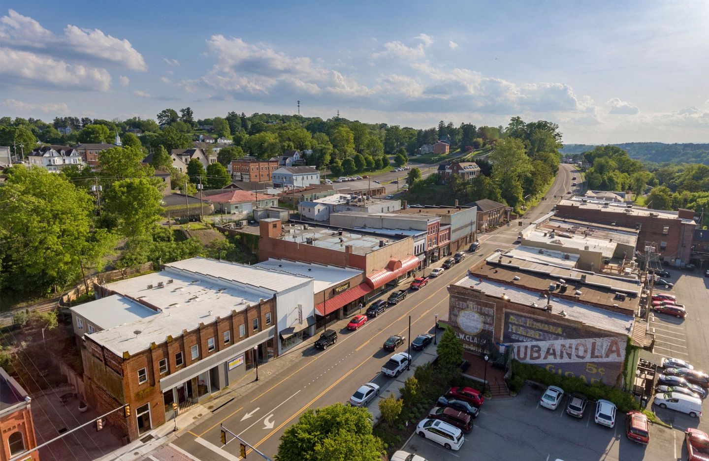 Aerial view of a small town main street with shops, parked cars, and tree-lined hills in the background