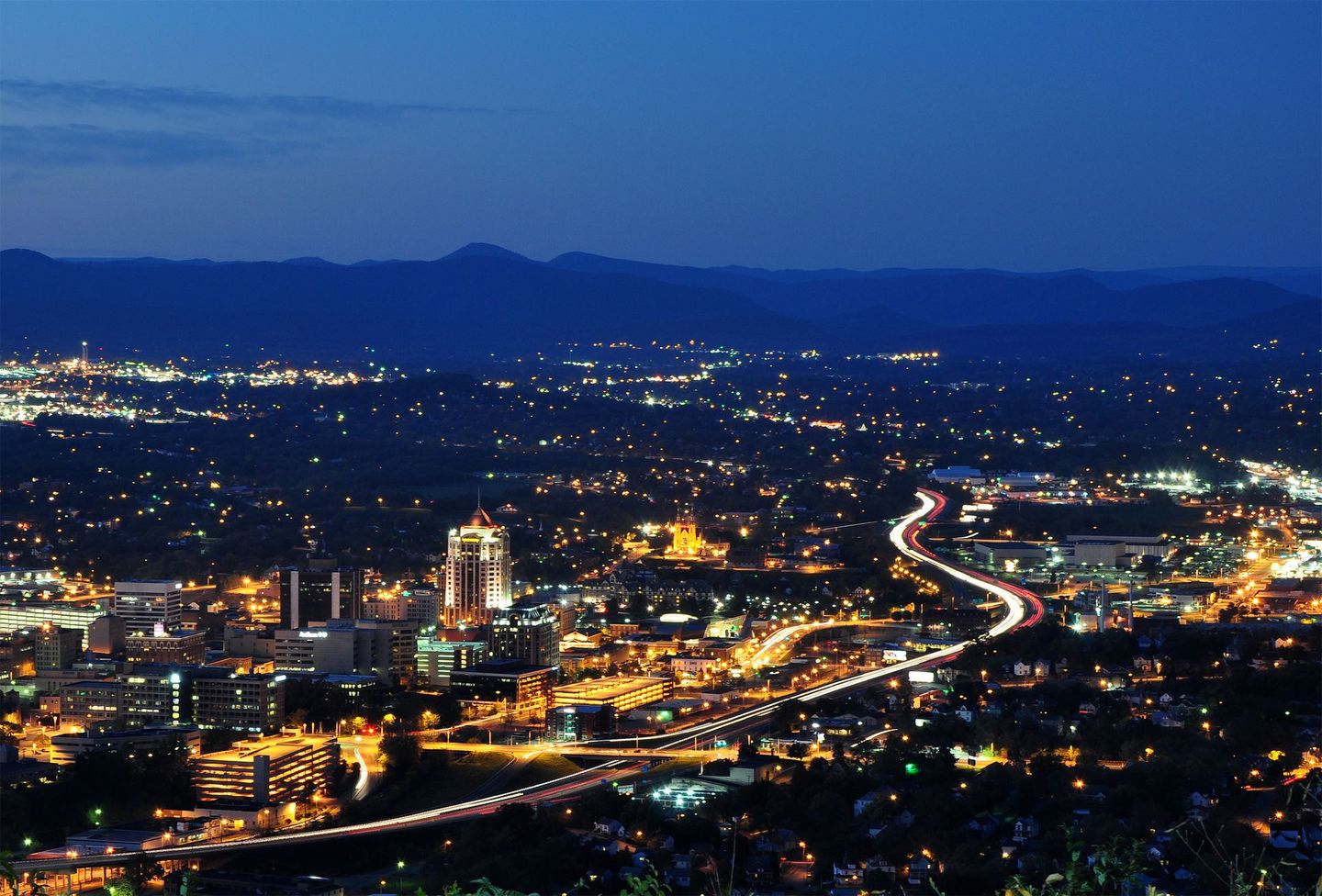Dusk cityscape of Roanoke with glowing lights and a winding highway stretching into the distance.