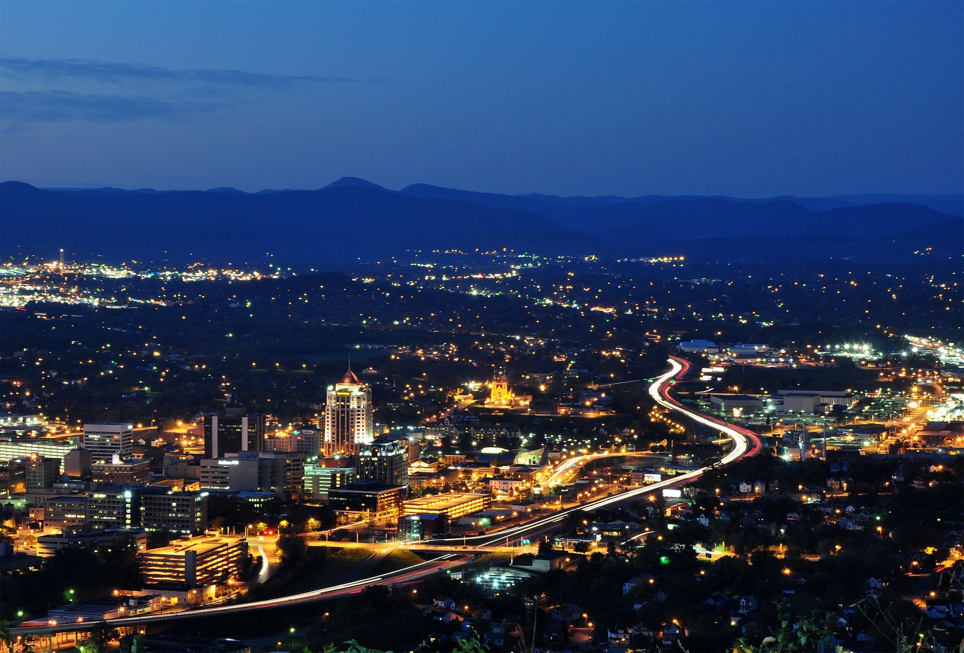 City lights and traffic trails at dusk, viewed from a hilltop over a sprawling valley and distant mountains in Roanoke, Virginia.