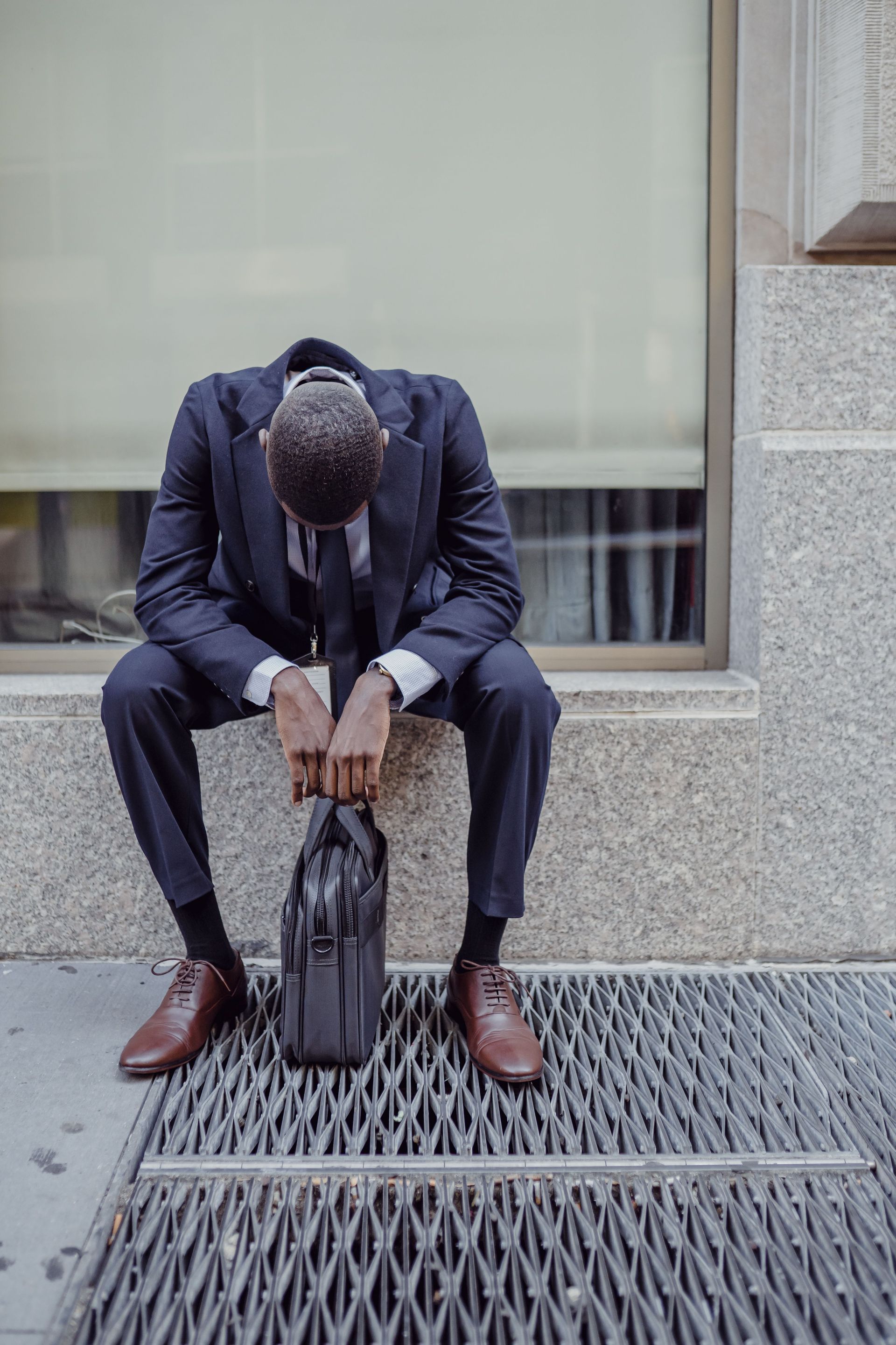 Person in a dark suit sits hunched on a ledge, head down, resting hands on a black bag.