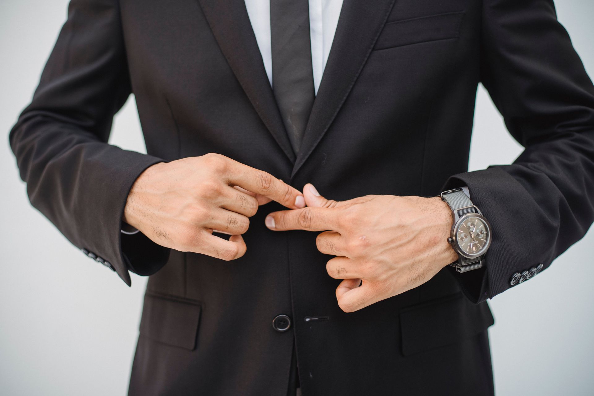 Person in a black suit fastening the front button of a jacket, wearing a wristwatch.