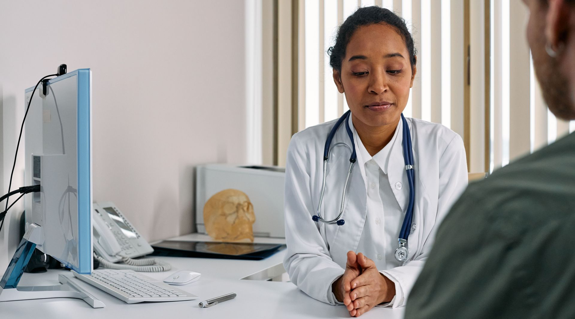 View our Service Area Doctor speaking with a patient in a bright medical office, seated at a desk with a computer.