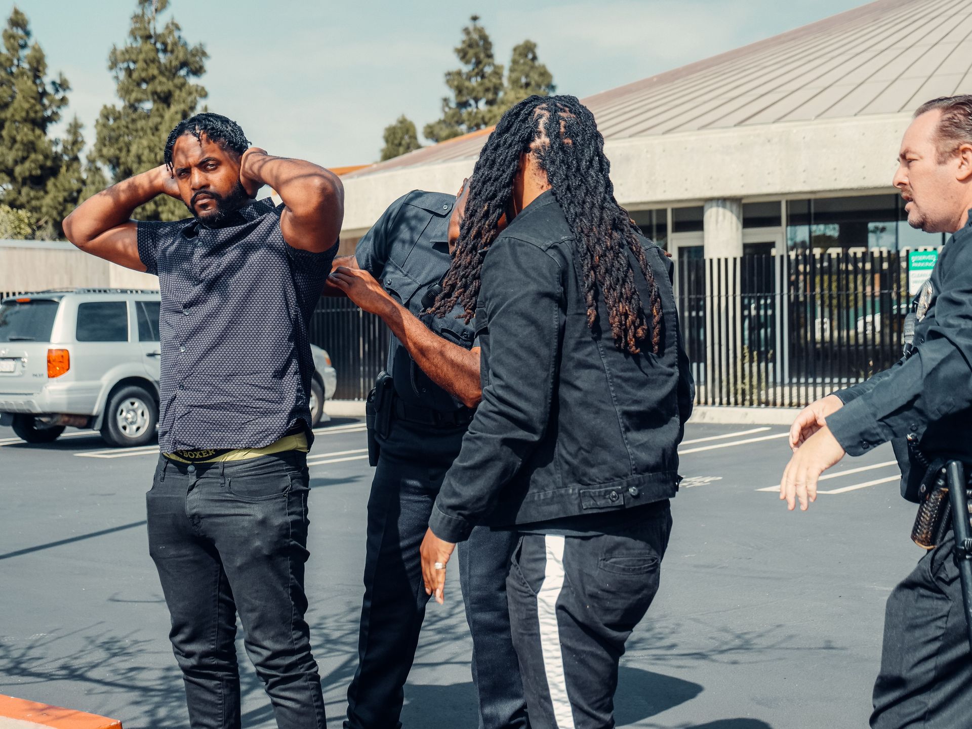 Three people standing in a parking lot, one with hands on head and another reaching toward them