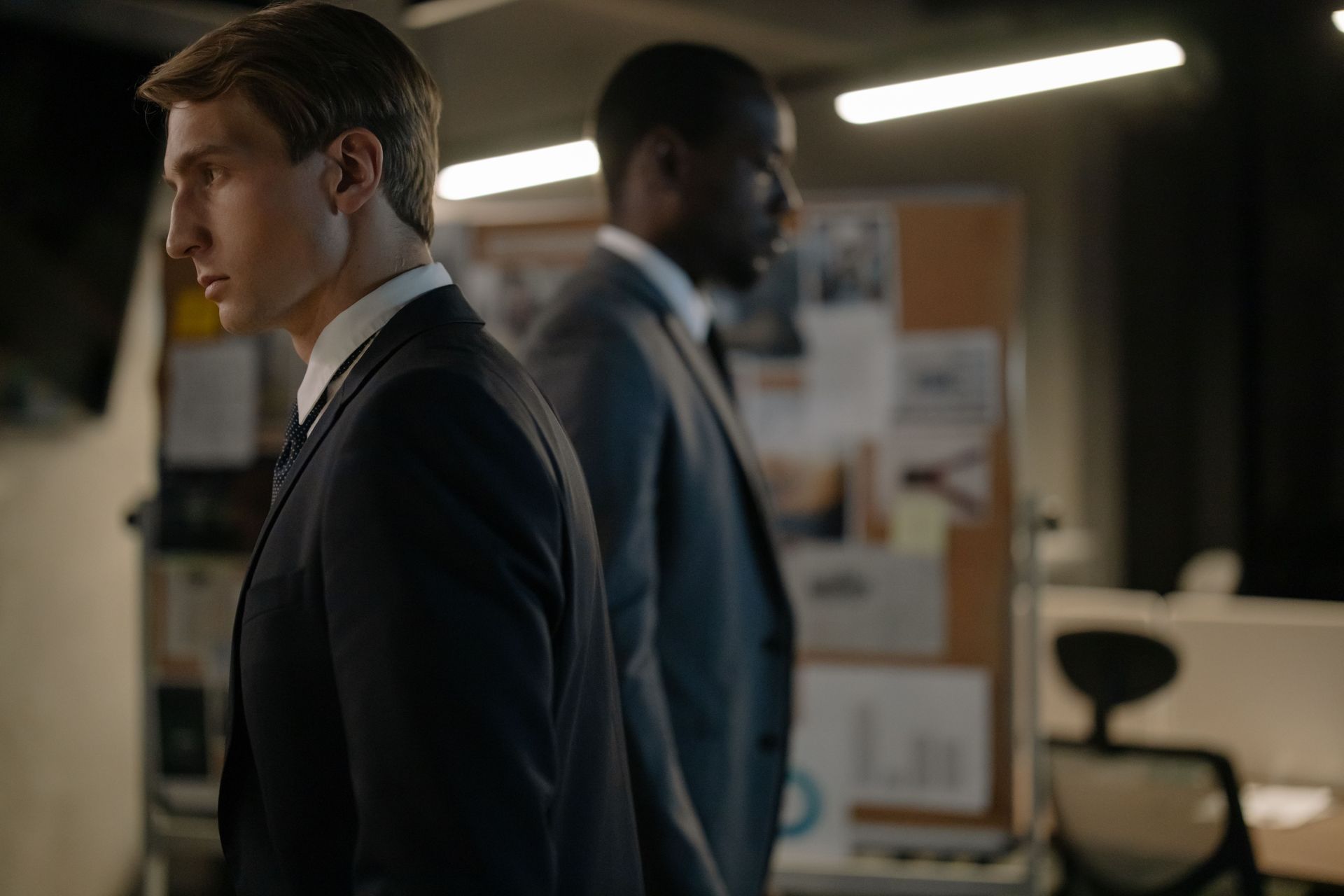 Two men in suits standing in a dim office, one in profile near a bulletin board, the other blurred in the background