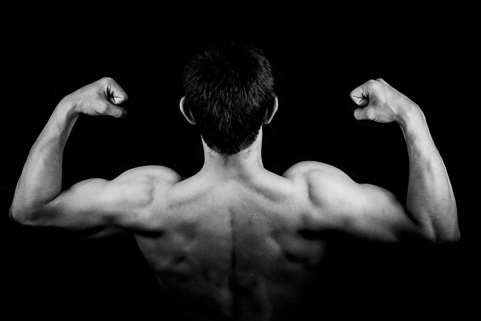 A muscular man flexes both arms, showing off his back and biceps in black and white.